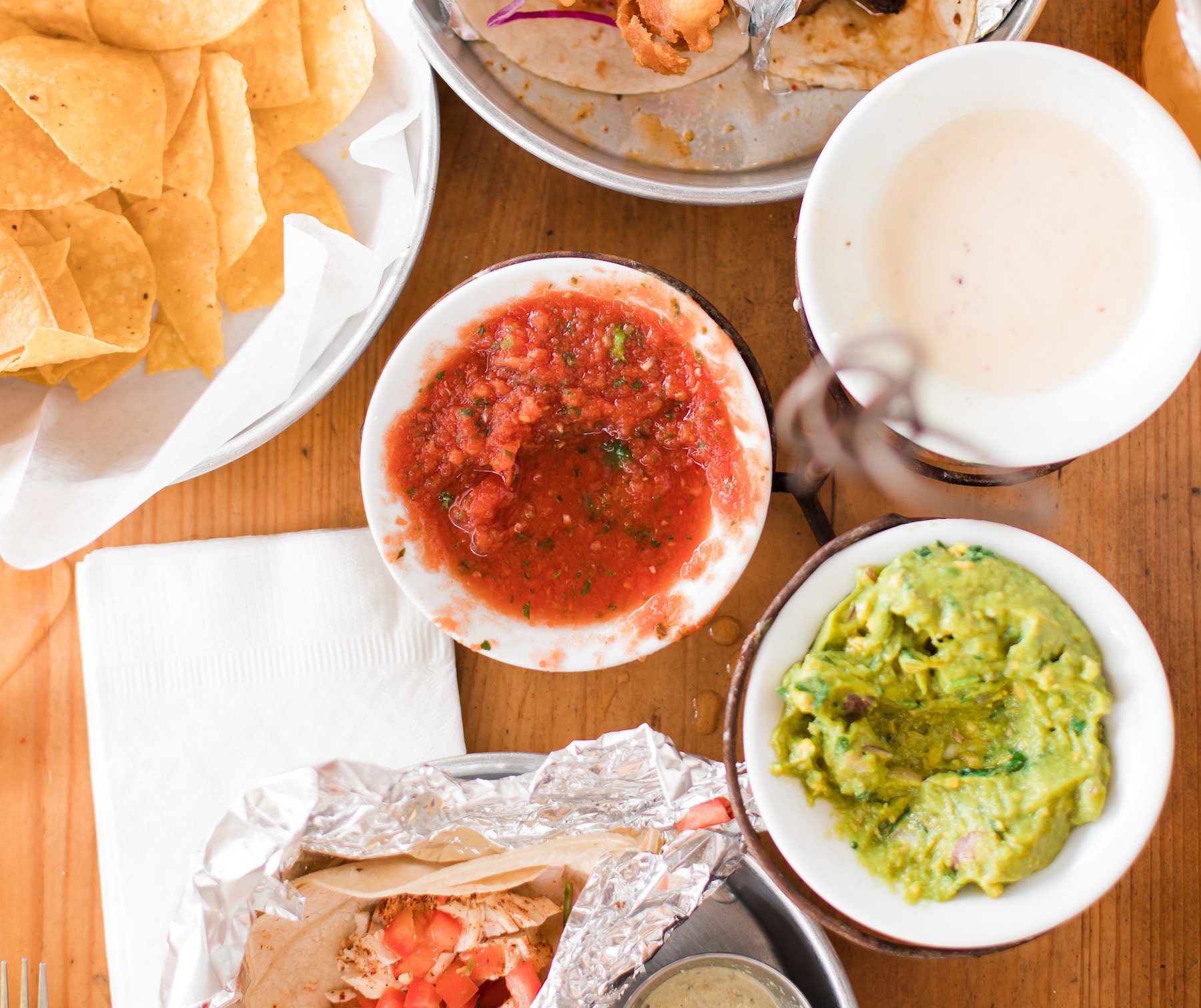 Wooden table filled with a plate of corn chips, burritos in foil, and dishes of salsa, queso, and guacamole. 