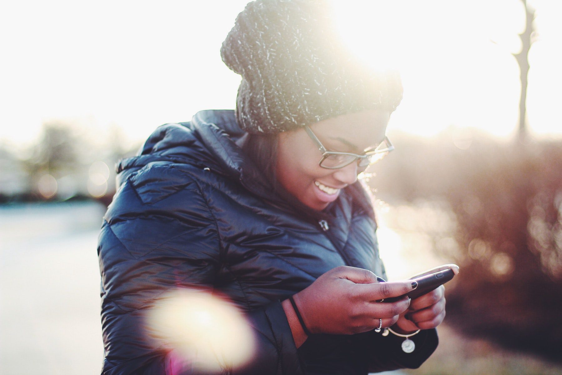 Young Black woman in winter coat and slouchy knit hat smiling at her phone with sunlight glare behind her. 