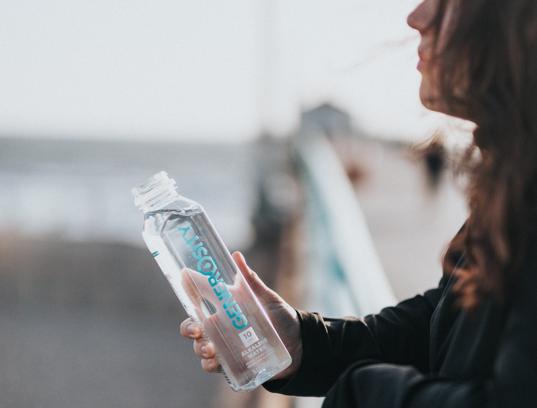 A dark-haired woman in a black sweatshirt holds a plastic water bottle while leaning on a bridge or pier.