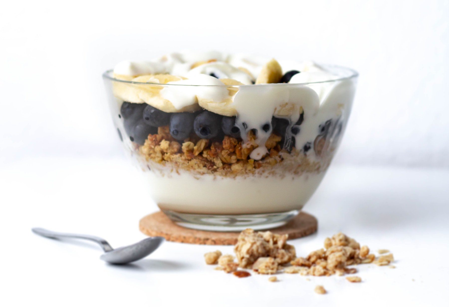 A clear glass dish of yogurt, blueberries, granola, and banana slices with a spoon nearby on a white counter.