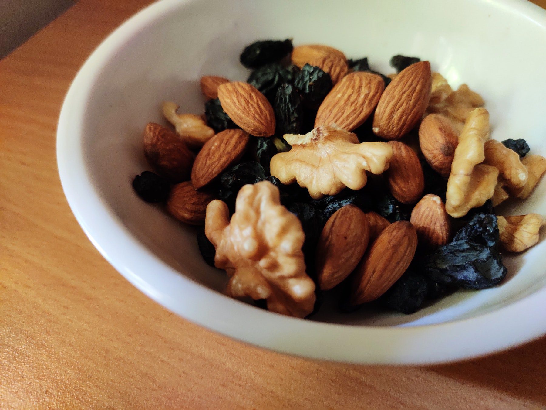 A white ceramic bowl of mixed nuts including almonds and walnuts on a wooden table. 