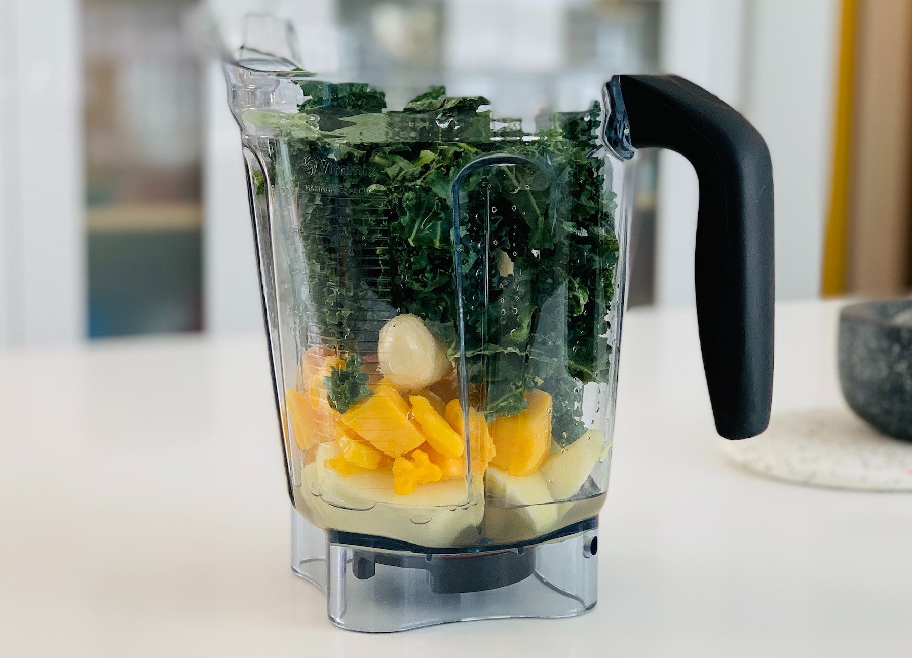 Ingredients for a smoothie recipe piled into a blender pitcher, including mango, banana, and kale.