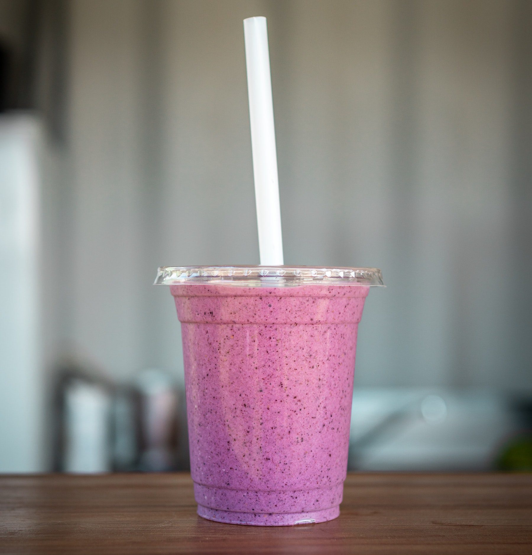 A purple-hued berry fruit smoothie in a clear cup with a tall straw on a wooden counter.