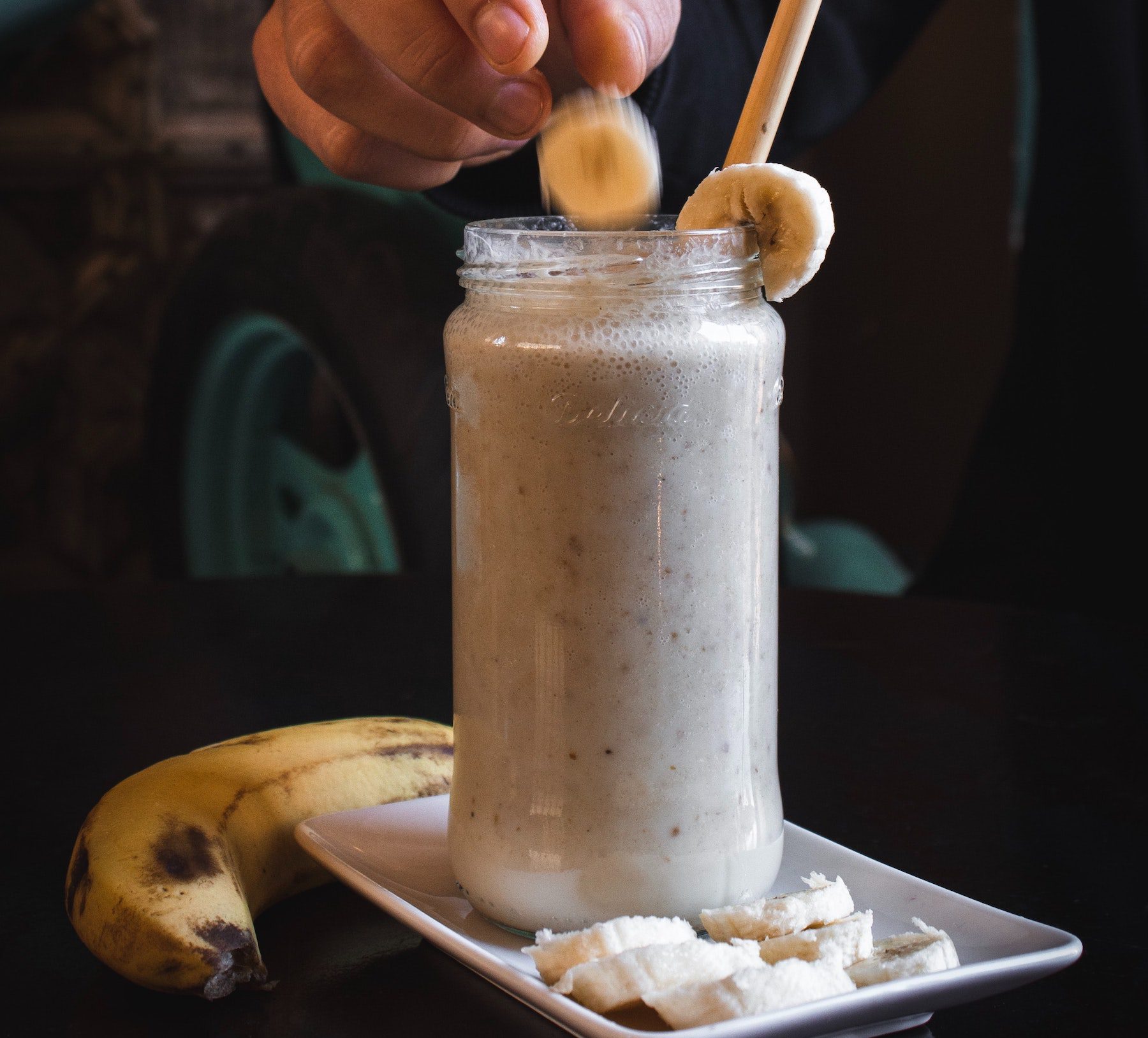A tan-colored smoothie in a clear jar being topped with banana slices, on a ceramic tray with more bananas.