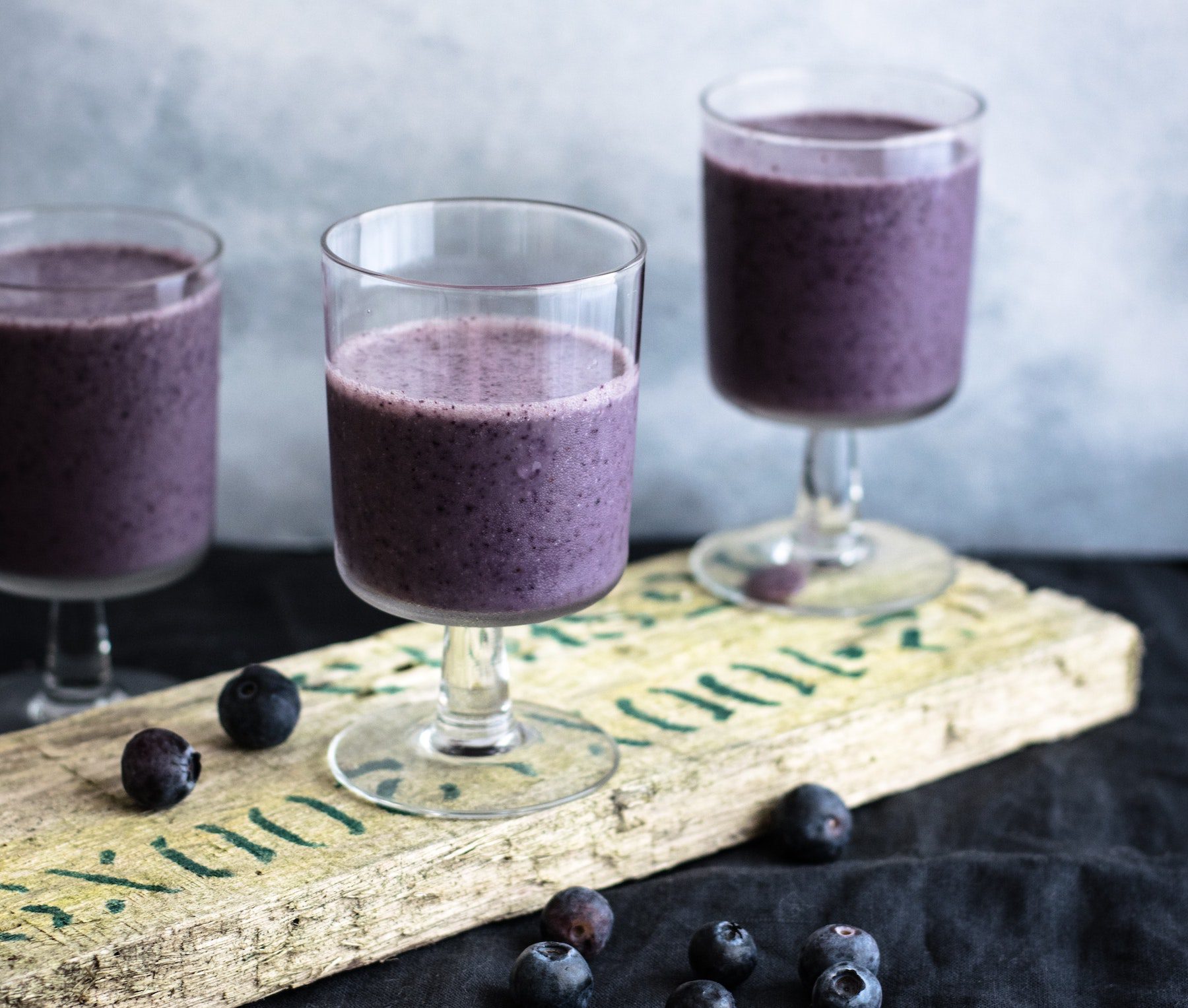 Blueberry smoothies in small decorative glasses on wooden serving tray, with fresh berries scattered nearby.