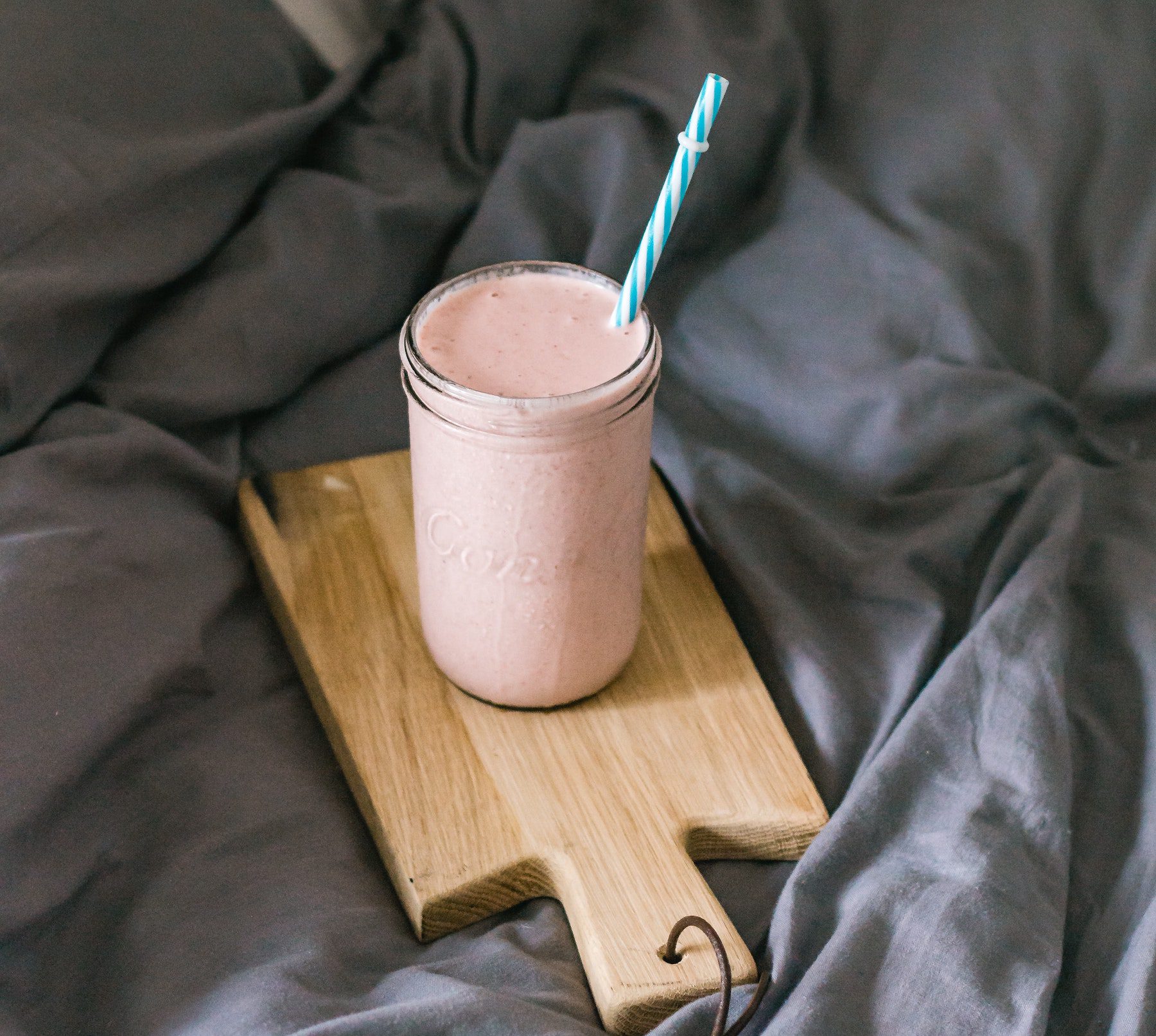 A classic strawberry smoothie in clear jar and blue paper straw on a wooden cutting board.