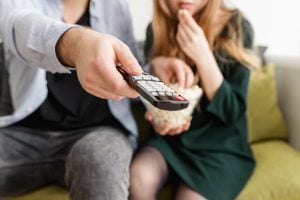 A man and woman sitting on a gray couch eating popcorn and scrolling TV shows with remote.