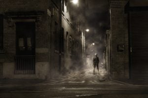 A creepy lone figure stands in a dimly lit, foggy alley.