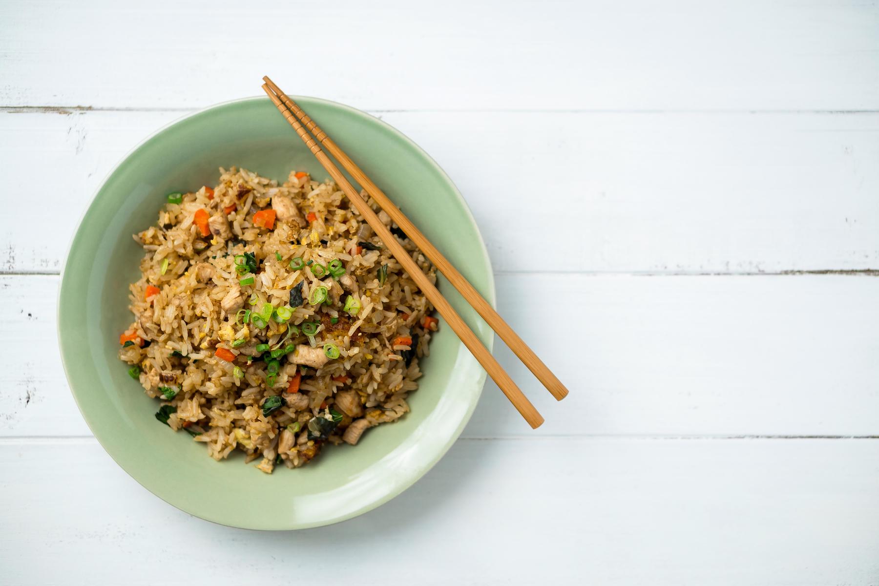 Fried rice with vegetables in a light green bowl on a white plank table with chopsticks.