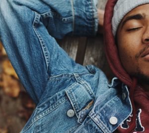 Black man in jean jacket and maroon hoodie naps on a park bench during autumn.