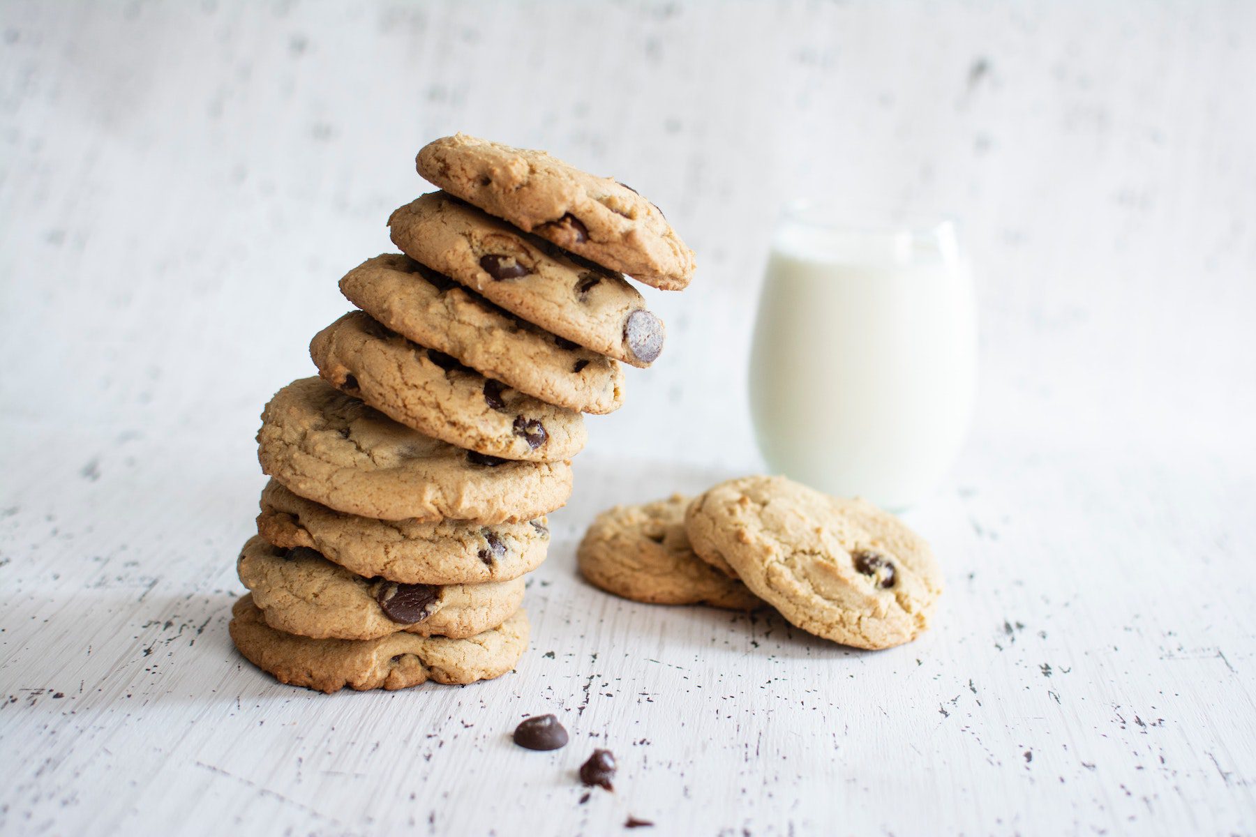 A leaning tower of vegetarian chocolate chip cookies on speckled background with a glass of milk nearby.