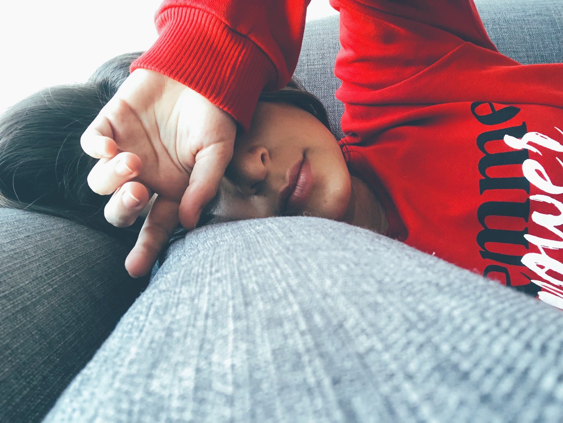 Young brunette woman in bright red hoodie naps with her head on a couch armrest. 