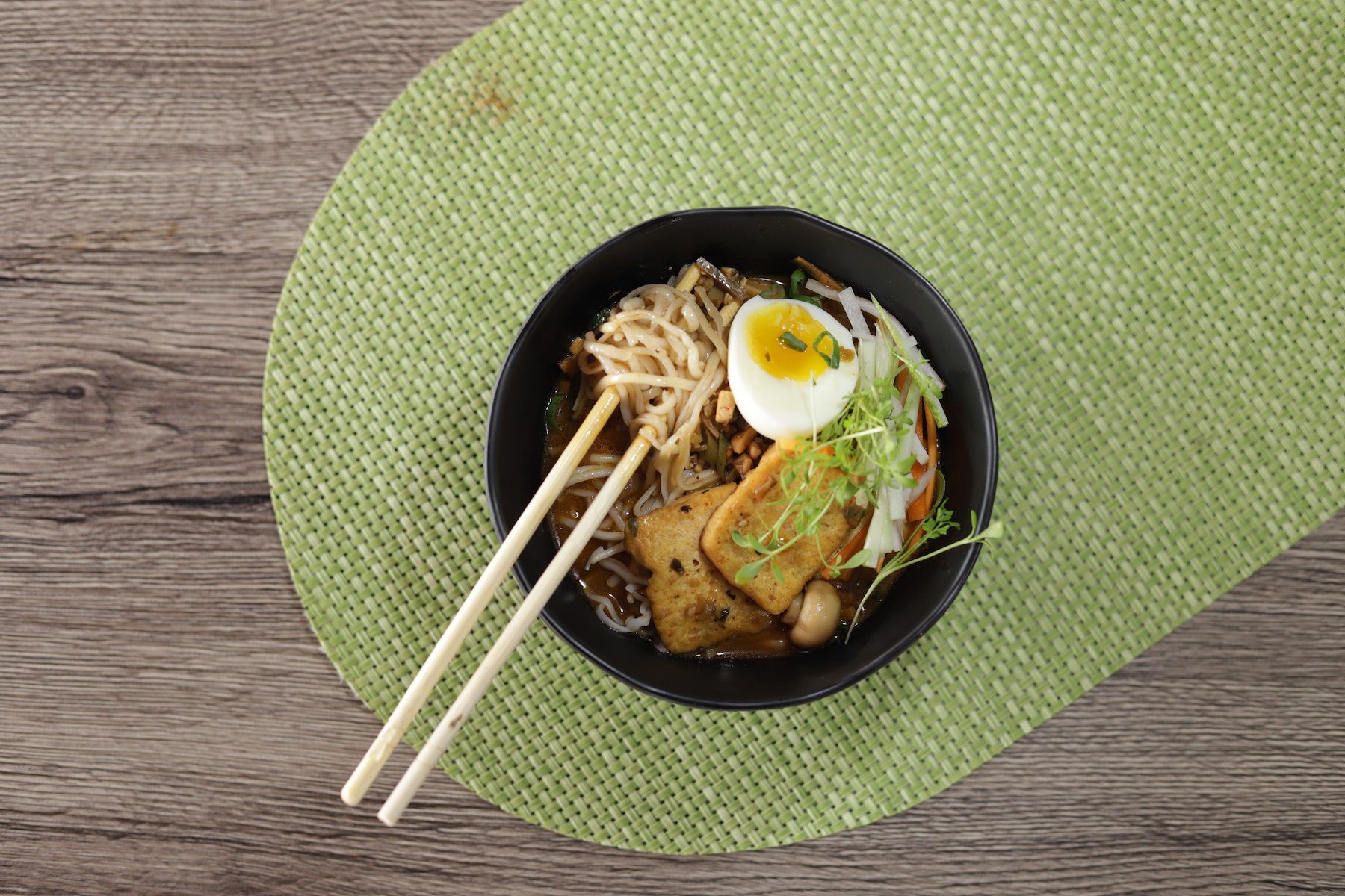 Bowl of ramen or other thin noodles with hardboiled egg, leeks, button mushrooms and tofu with chopsticks on woven green mat.