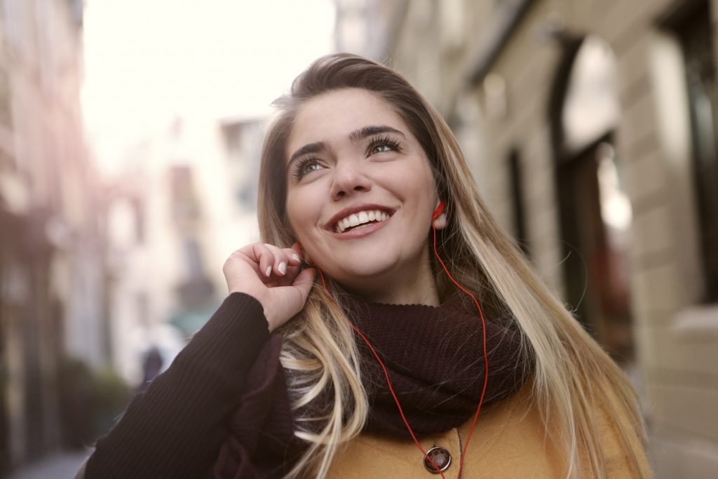 Beautiful blonde woman in brown scarf and yellow jumper listening to a podcast and smiling against blurred city backdrop.
