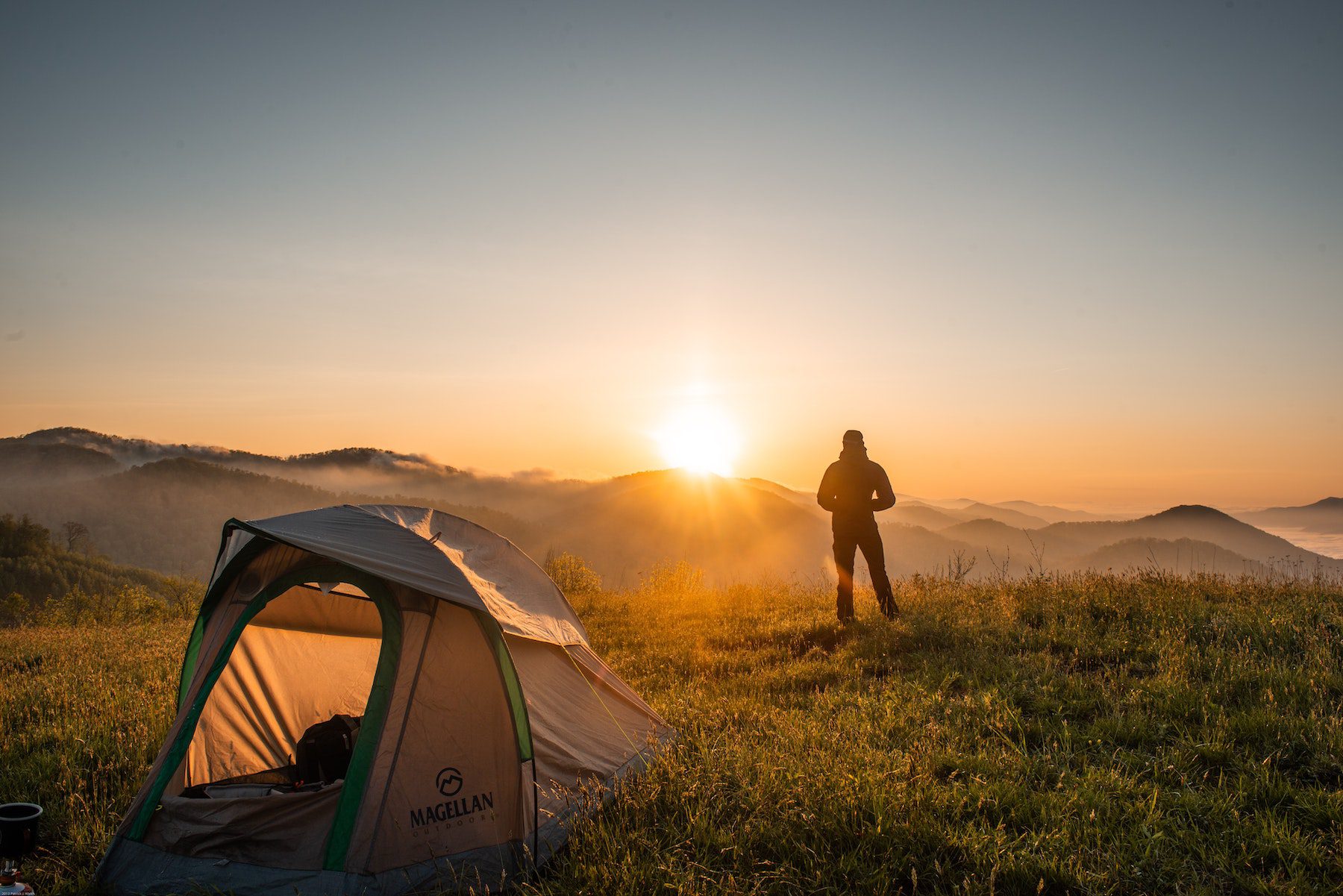A person stands and watches the sunrise near their tent on a grassy mountain top. 