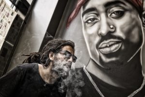 A Black man with dreadlocks and sunglasses exhales smoke while sitting in front of a mural depicted hip hop legend Tupac.
