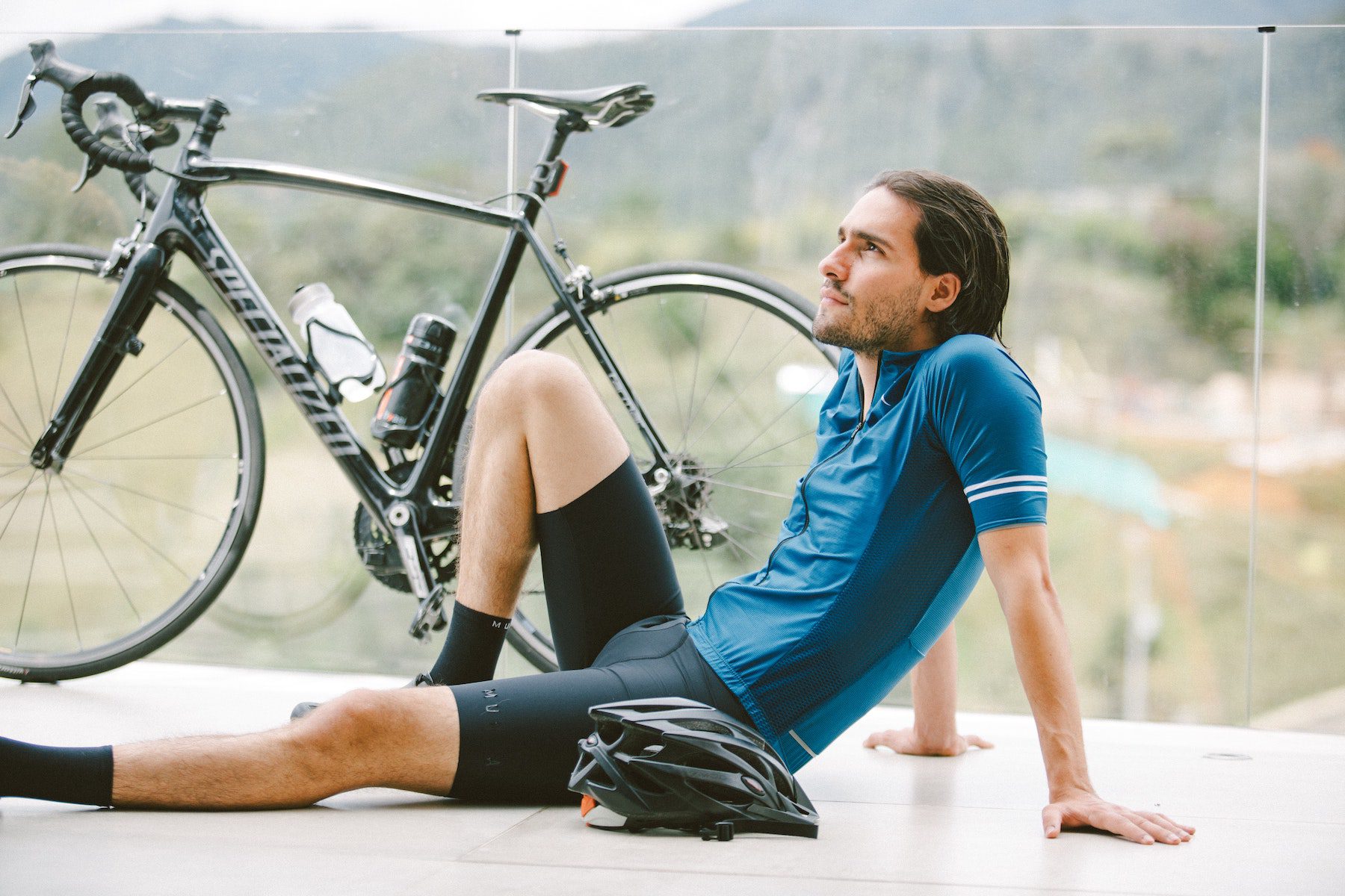 Cyclist in blue athletic clothing and black cycling shorts, resting on a glass-lined balcony beside a black bike.