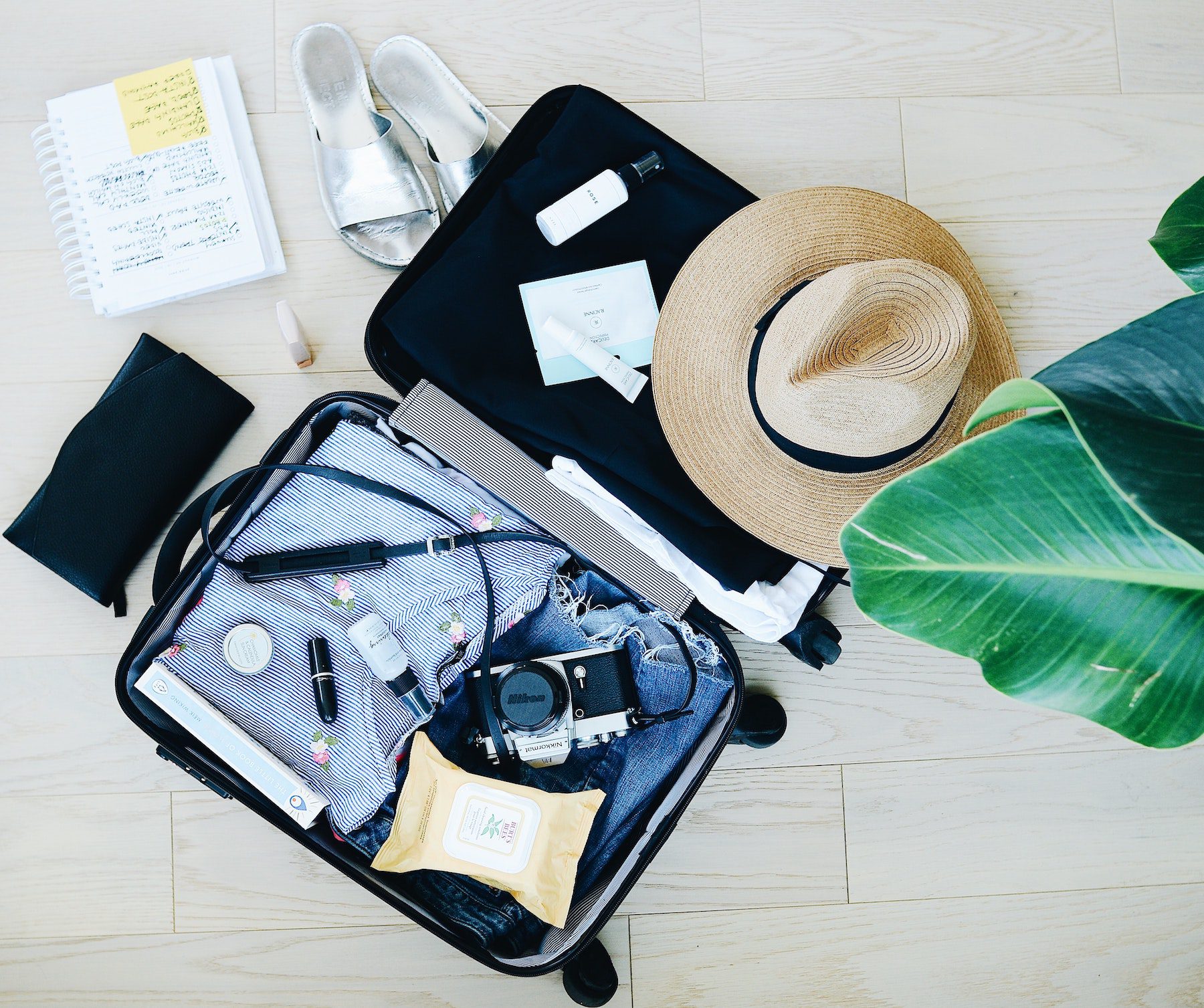 Suitcase flatlay featuring hat, jeans, camera, pen and paper, shoes, and other travel necessities. 