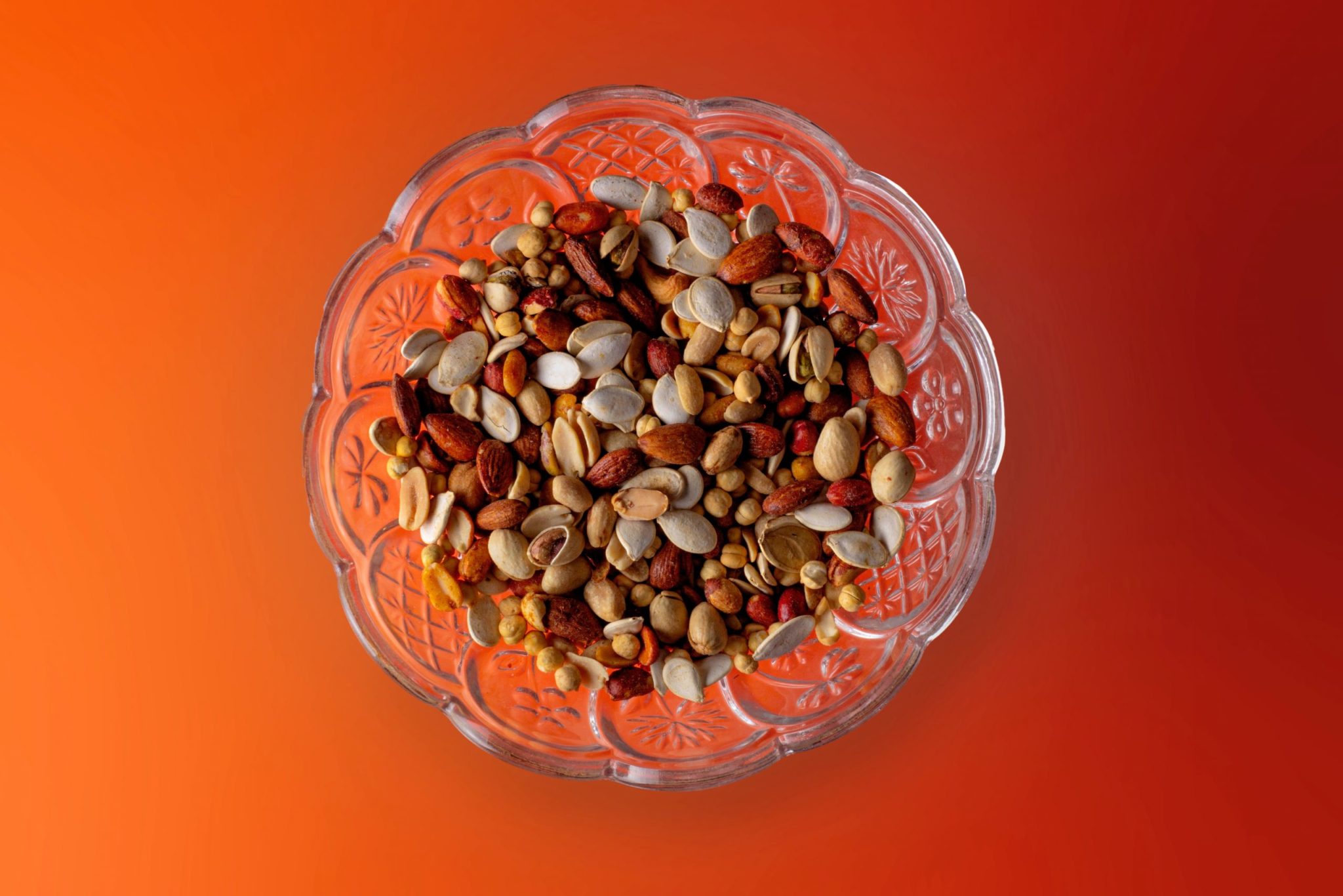 An ornate glass bowl filled with a variety of nuts on an orange table.