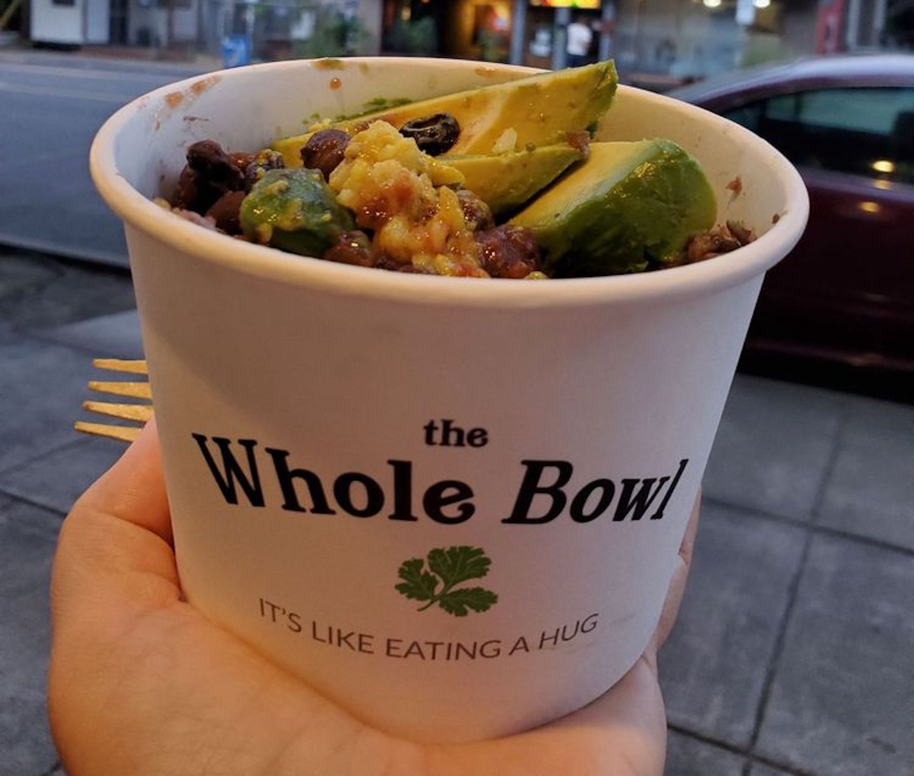 Person holding paper Whole Bowl cup that reads "Like Eating a Hug," filled with avocado, black beans, and other vegetarian ingredients.