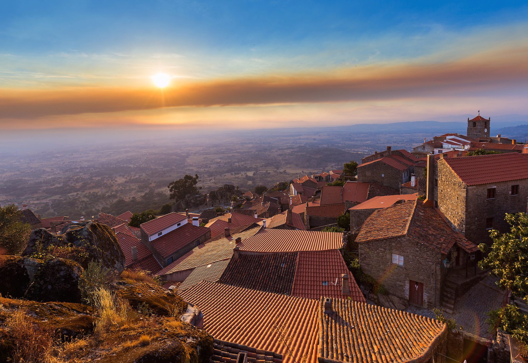 Sweeping hilltop view of Monsanto, Portugal were the sun is beginning to set behind wisps of hazy clouds