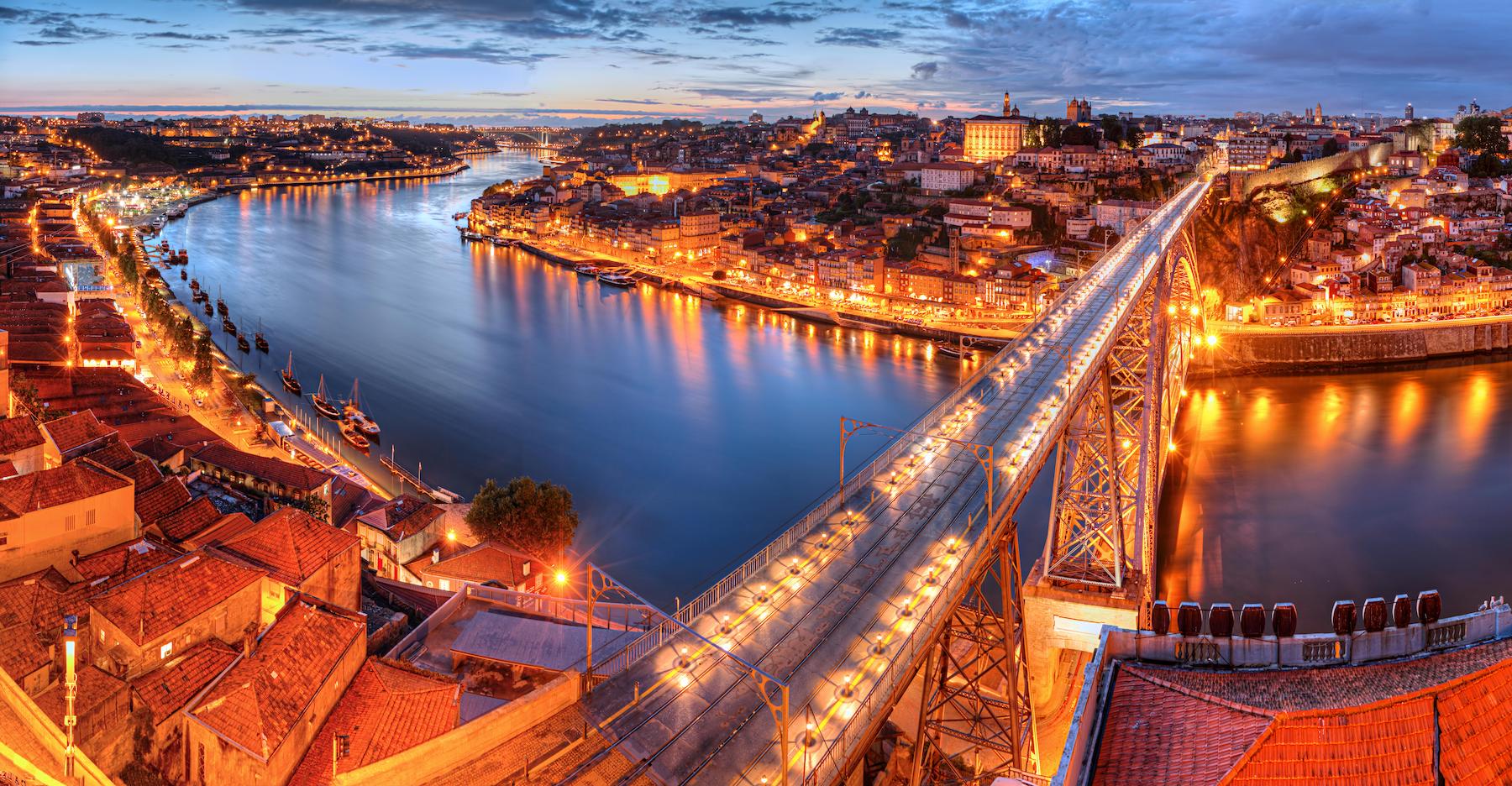 Ribeira District in Portugal on partly cloudy evening as the streetlights from the city light up the river under the bridge
