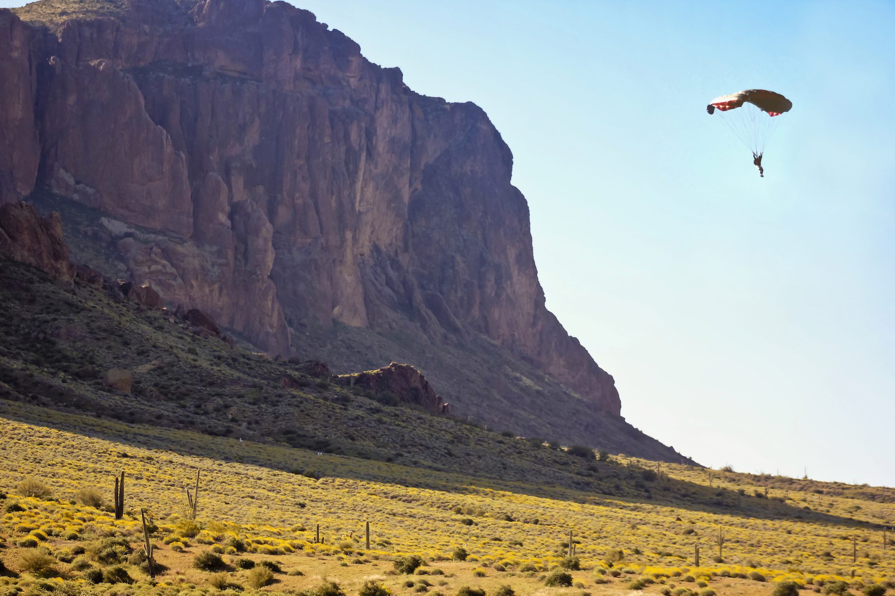 A paraglider is shown approaching landing in an open flat plain with a soaring cliff face in the background at Lost Dutchman State Park
