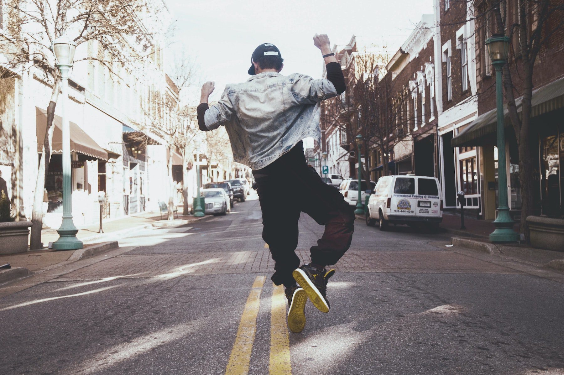 A man wearing a jean jacket and dark pants dancing in the middle of the street by clicking his heels together and arms raiseed
