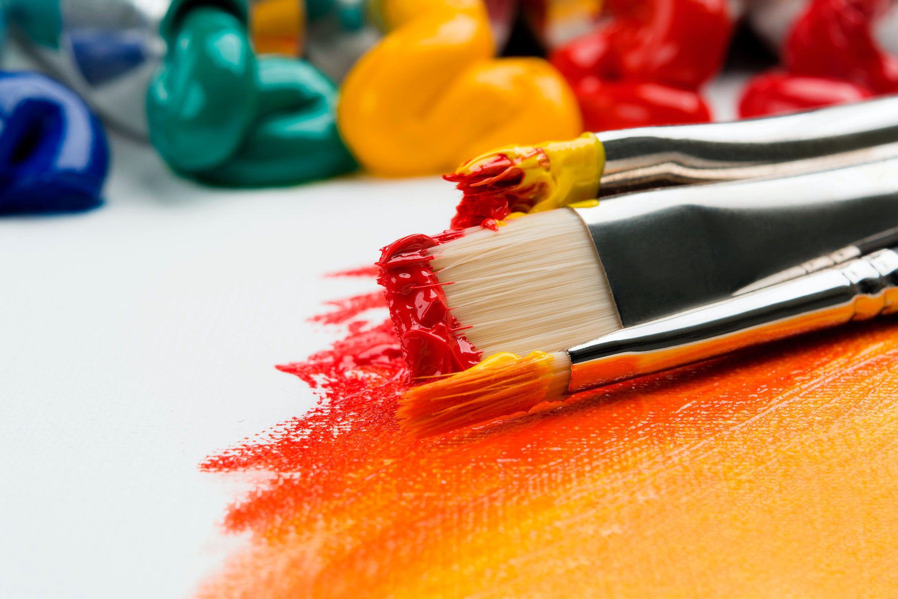 Paint brushes that were dipped in red and orange lay on a white canvas with various other colors blurred in the background