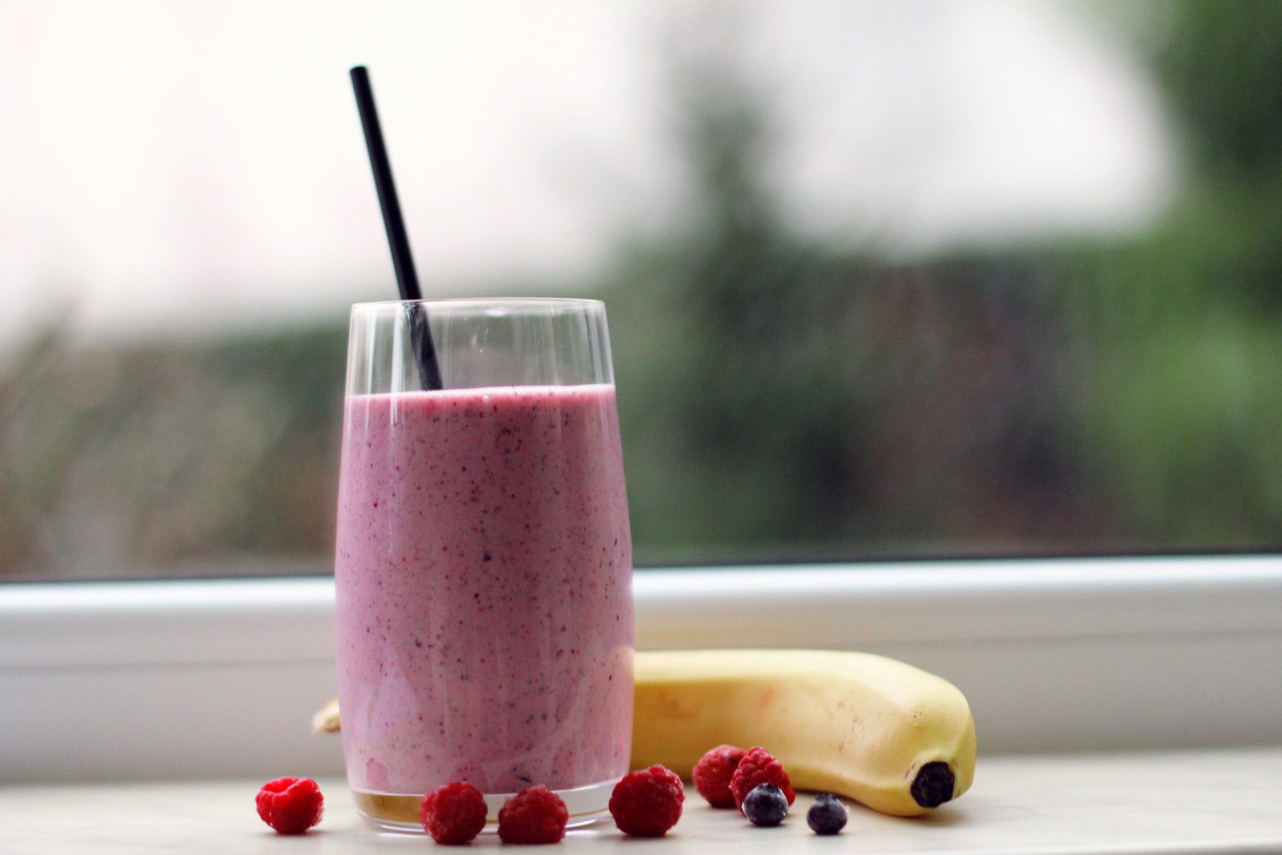 Pink, berry smoothie in a glass with black straw sitting in front of a window with a banana, raspberries and blueberries