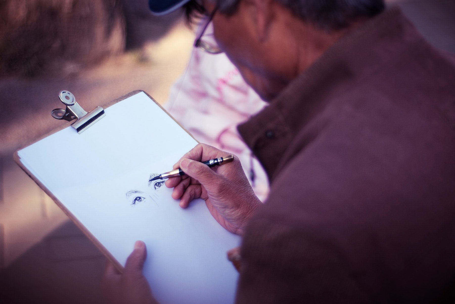 An artist wearing glasses, a hat and a brown coat sketching a pair of eyes on a blank sheet of paper on a clipboard