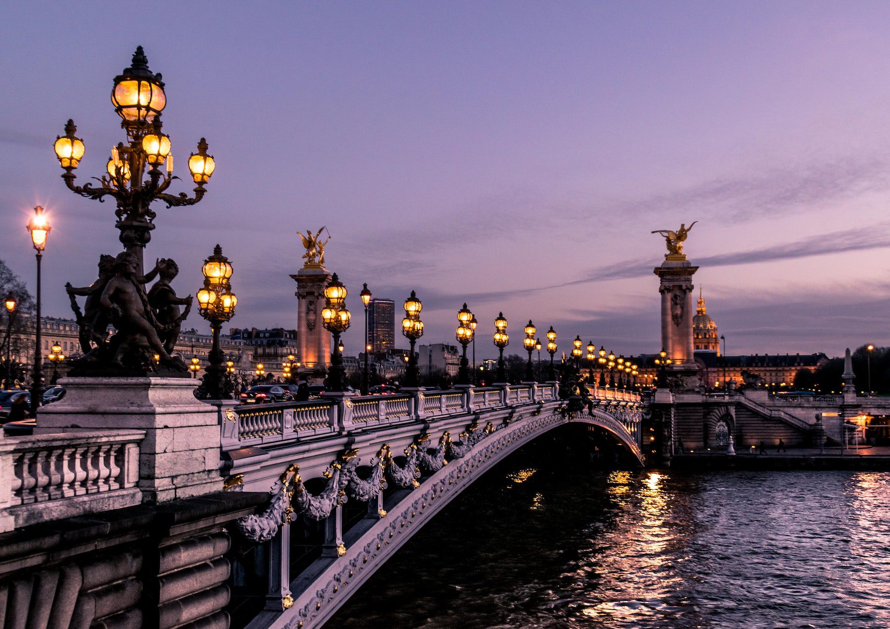 An antique style street-lamp lit white bridge extends across the water on an evening in Paris