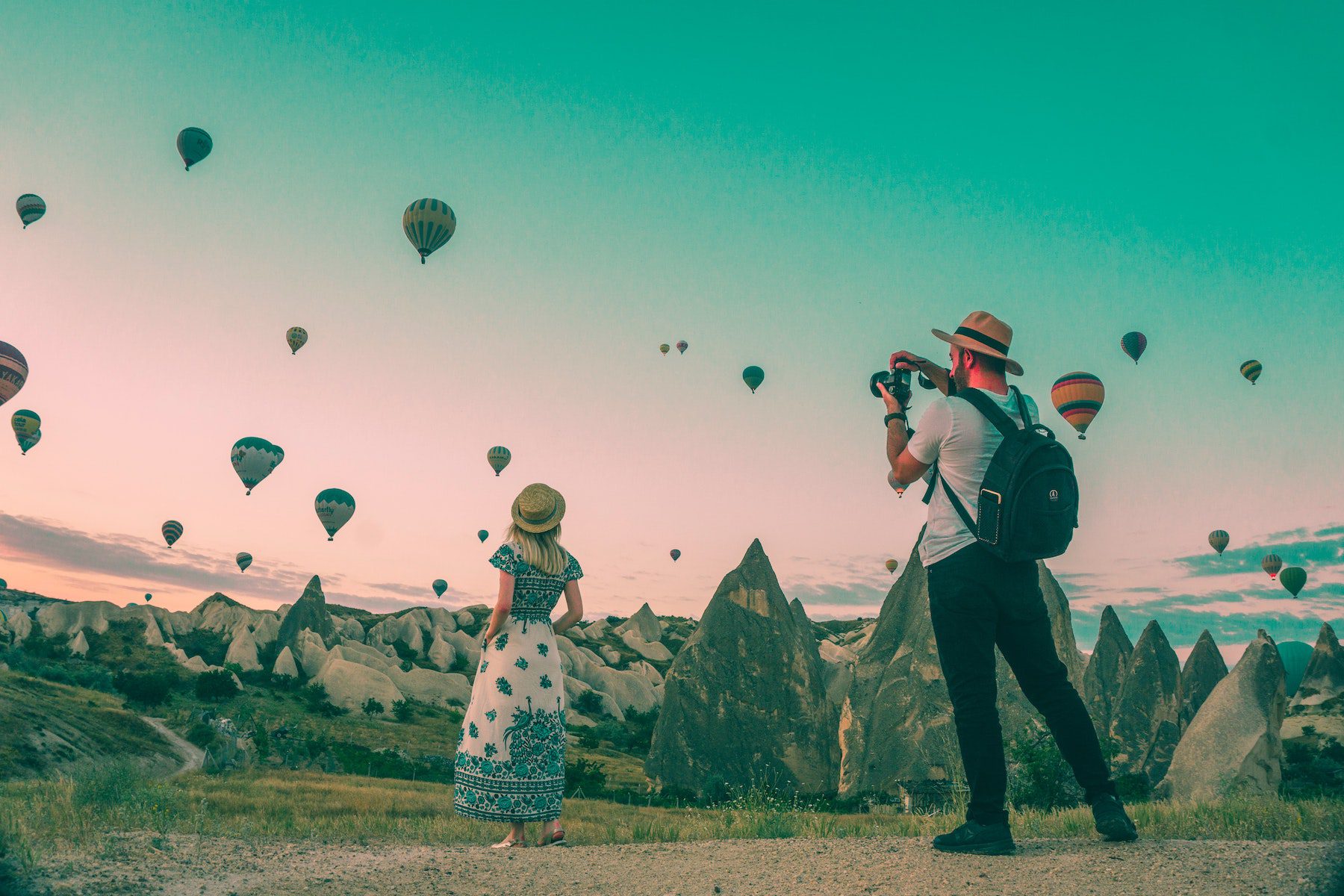 A man wearing a fedora, white t-shirt, and dark pants, photographs a woman in a blue and white dress as countless hot air balloons litter the sky above small sharp peaks