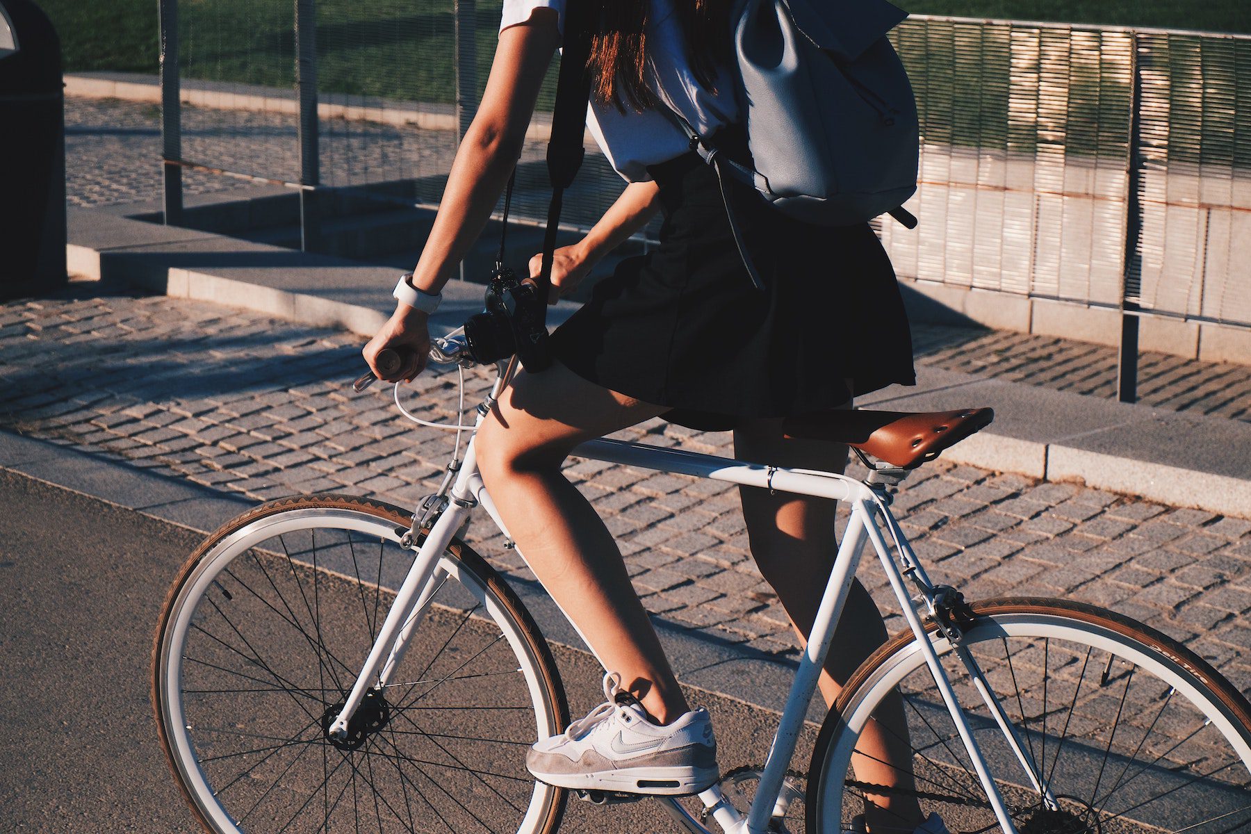 A girl in a black skirt and whit shirt wearing a small blue backpack rides a white bicycle on a paved road alongside a brick sidewalk 