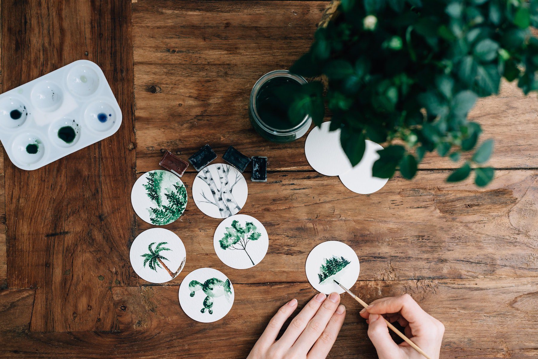 An artist paints plants, trees, and other greenery on coaster sized canvases atop a large wooden table