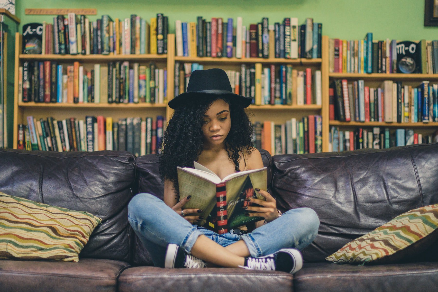 A girl wearing a black fedora and blue jeans reading a book on a dark leather couch with a small library of books behind her