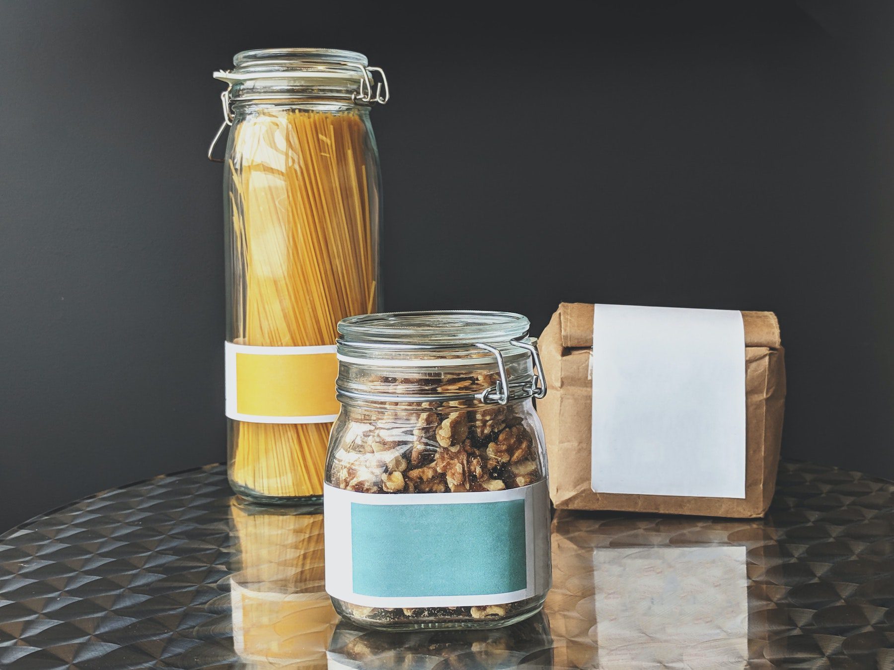 Uncooked pasta, walnuts are kept in separate glass jars next to a folded brown paper bag on top of a metal table
