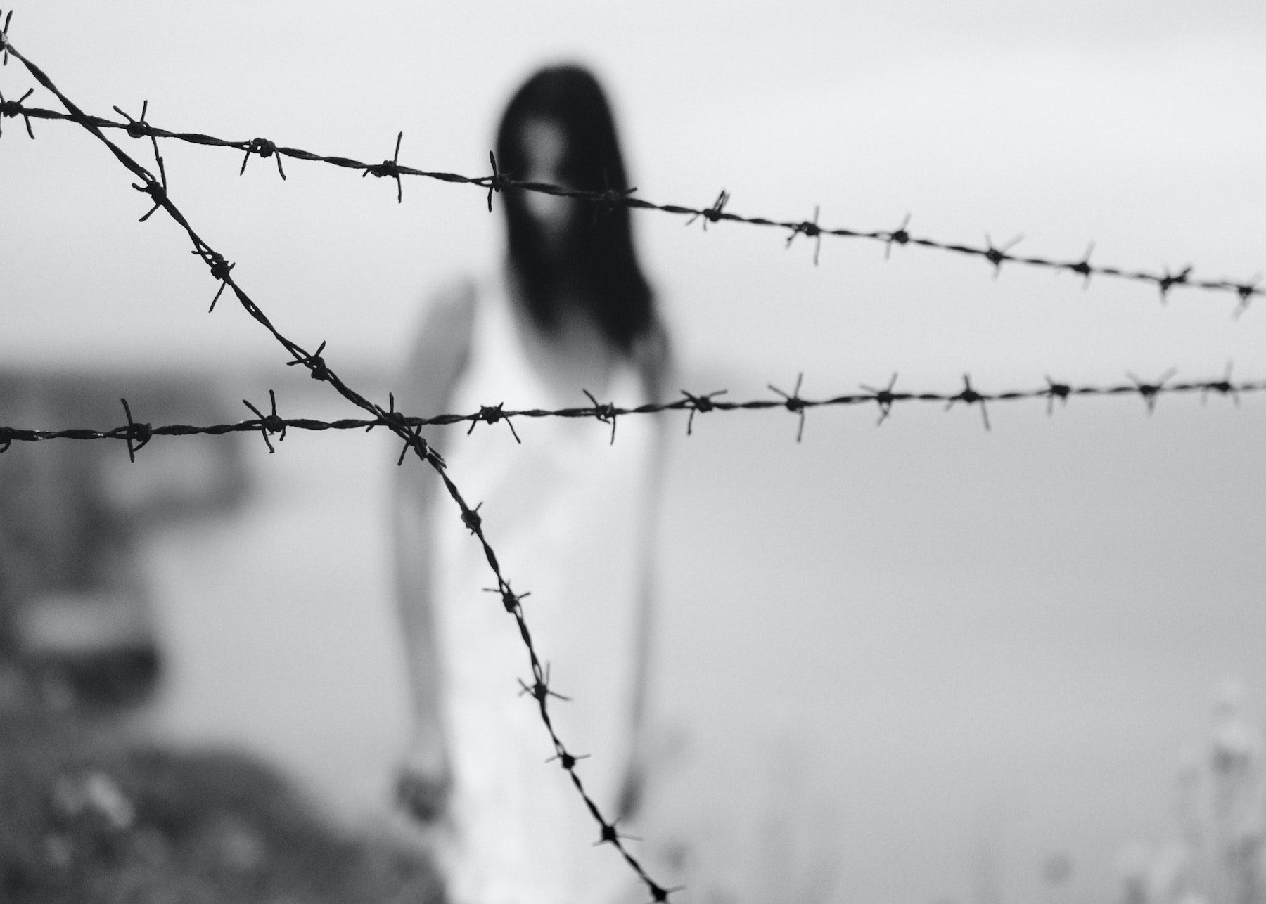 Blurred and eerie woman with dark hair in a white dress behind in-focus barbed wire.