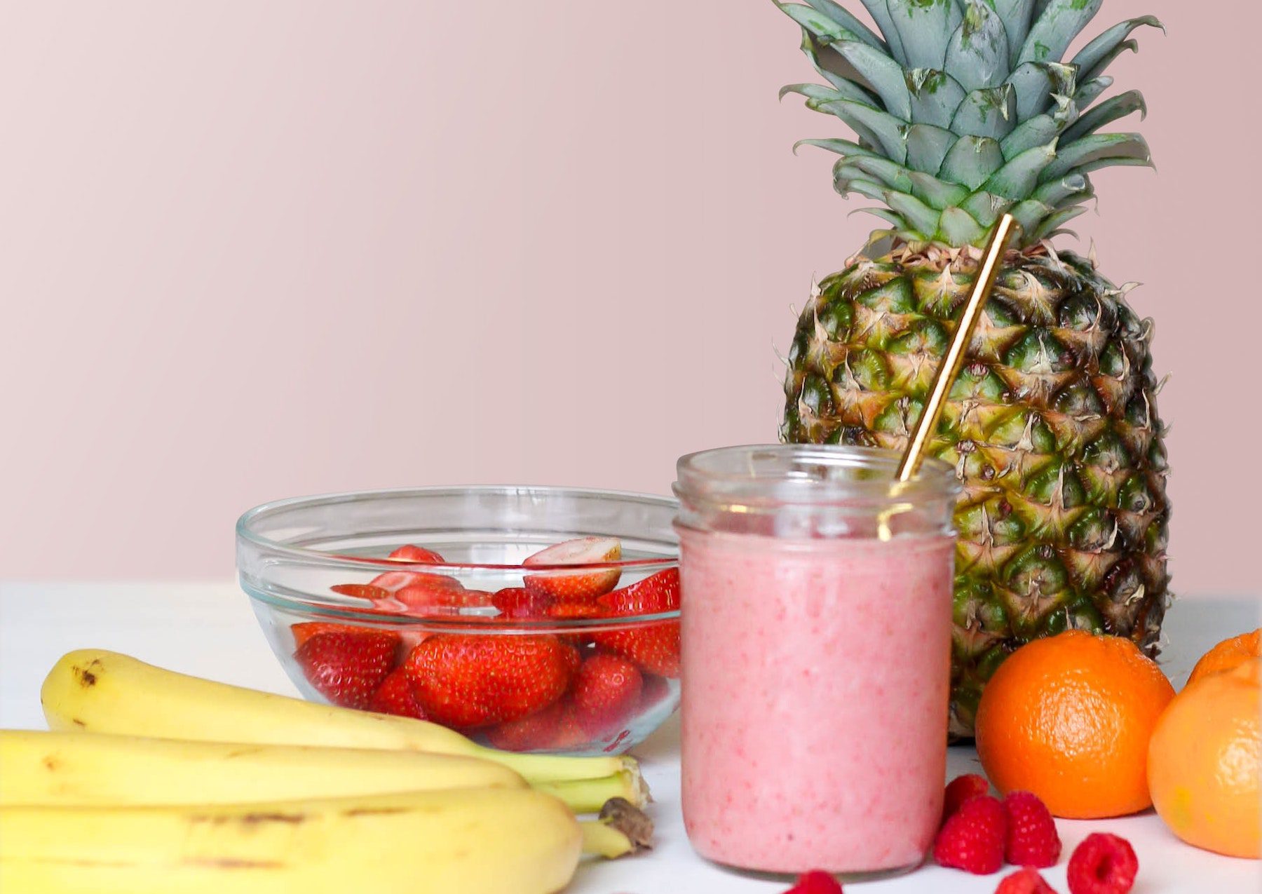 Pink smoothie in a mason jar in front of pineapple, bananas, strawberries, raspberries, and oranges against a pale pink wall.
