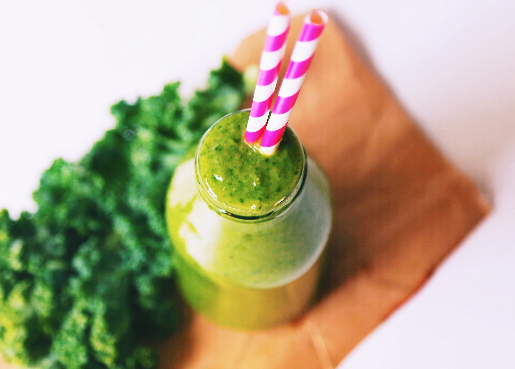A kale smoothie with fresh kale on a cutting board, with two pink paper straws inserted.