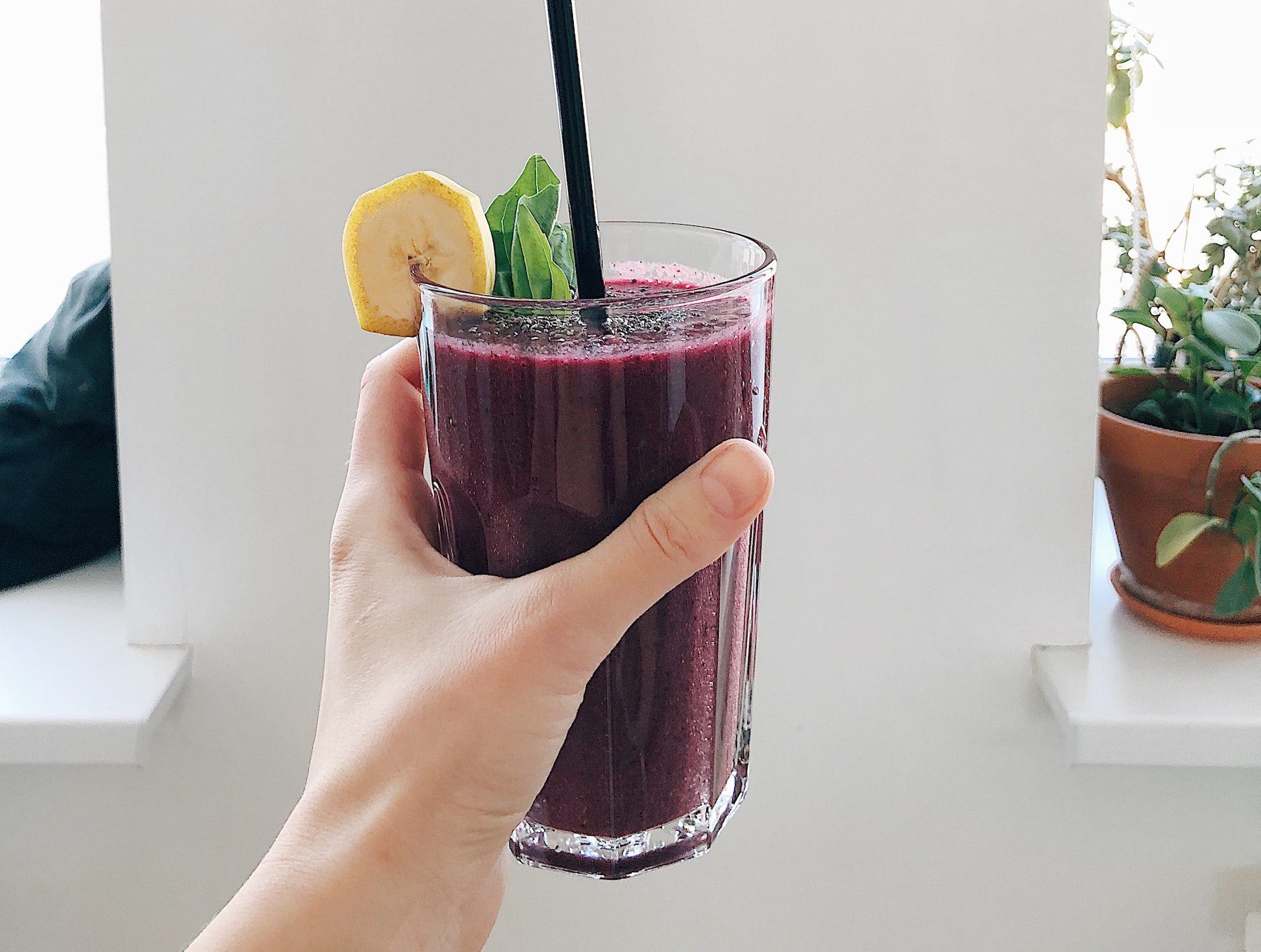 Hand holding a purple smoothie with a lemon wedge and fresh mint in front of a white wall.
