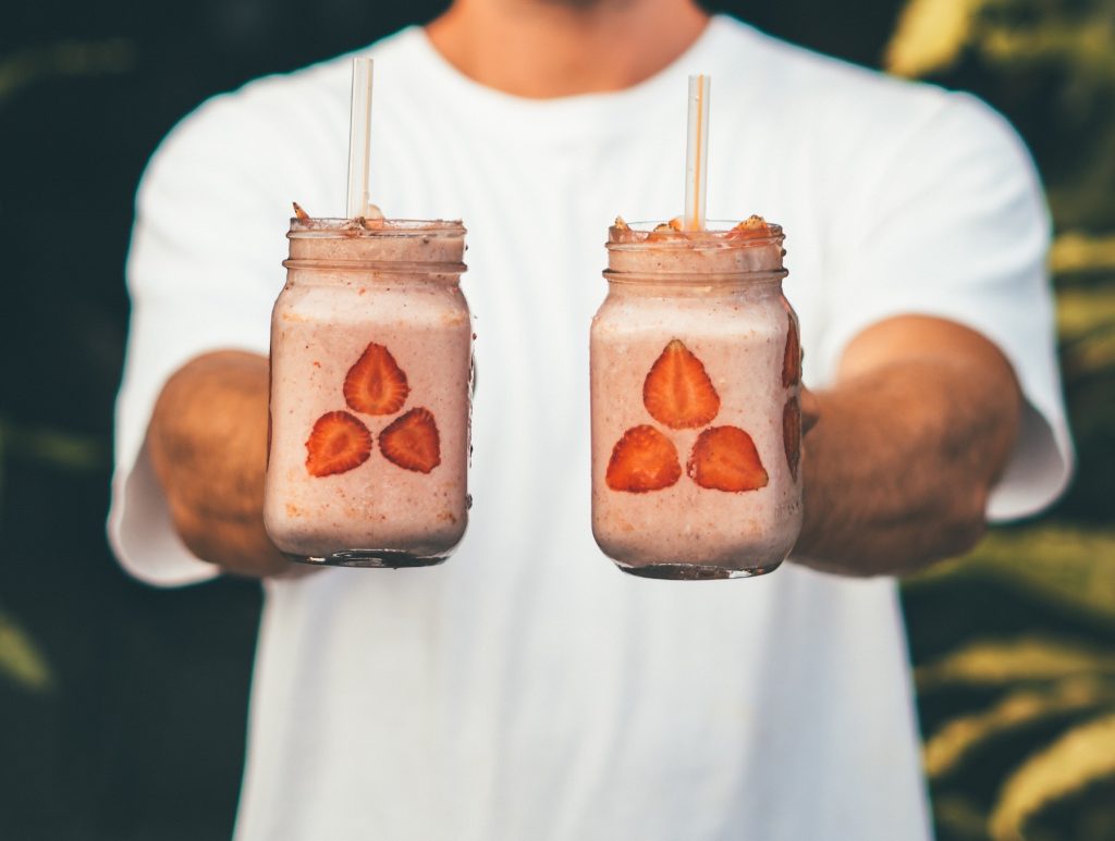 Man holding two mason jar glasses filled with strawberry smoothies with sliced strawberries against the glass.