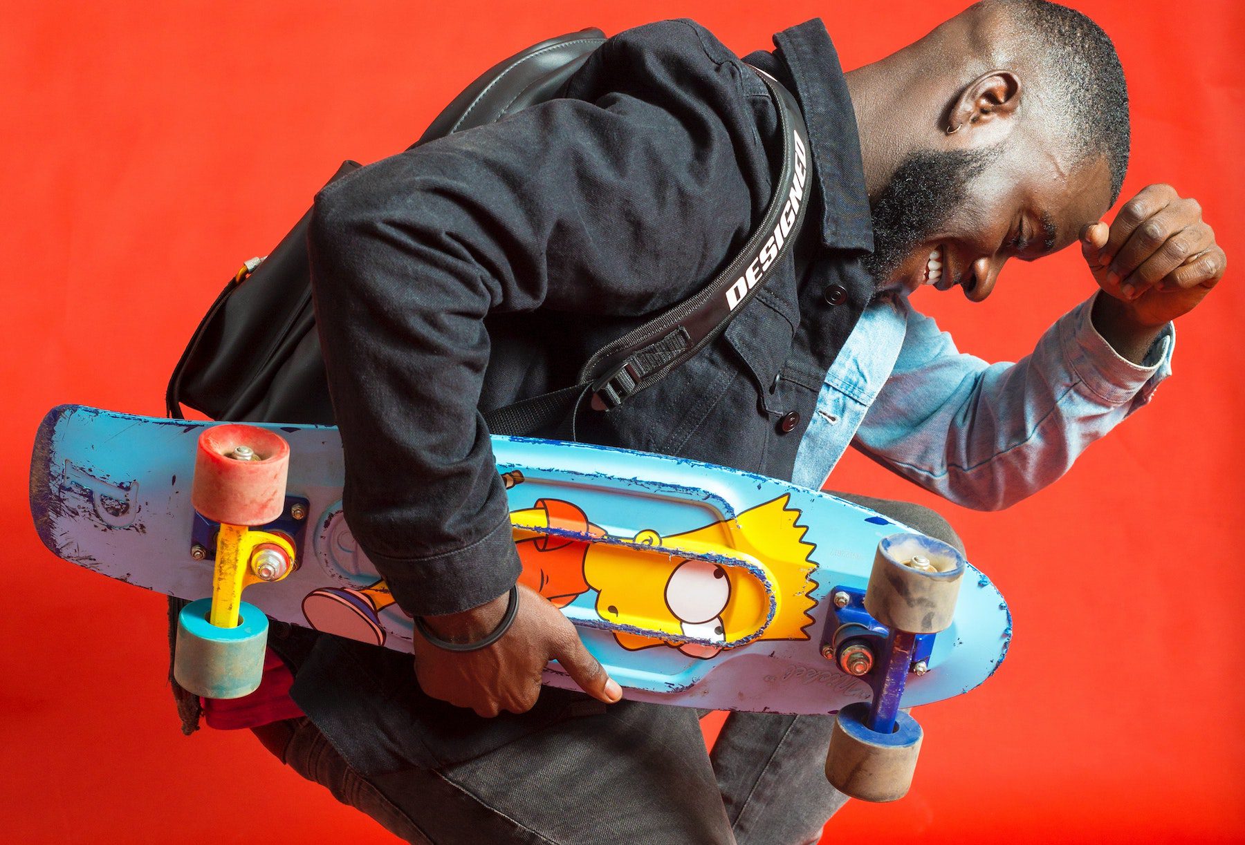 Young man holding a skateboard with Bart painted on the deck against a bright red-orange background.