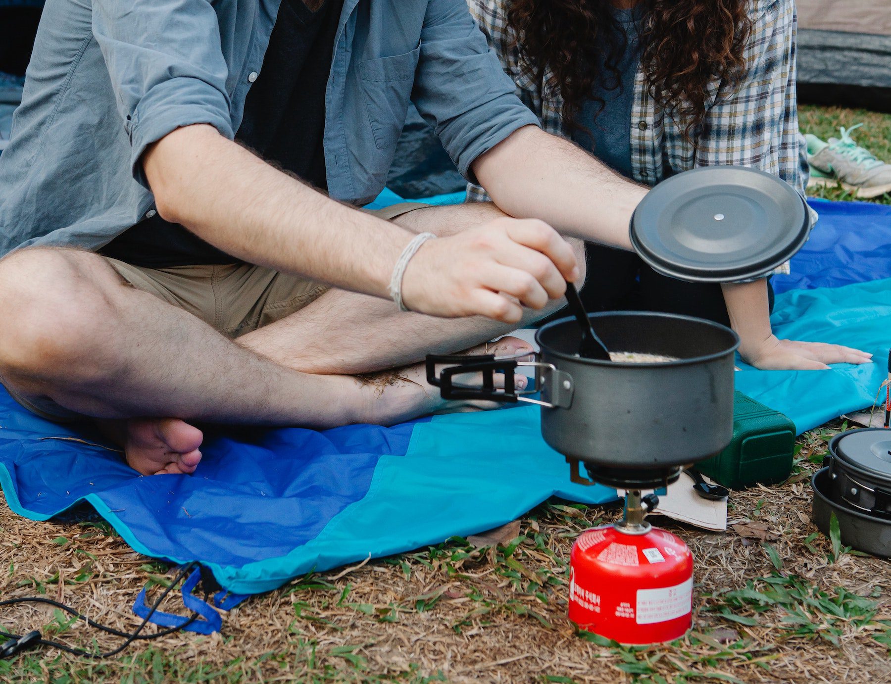 man stirring a meal inside a camping stove while sitting on the grass