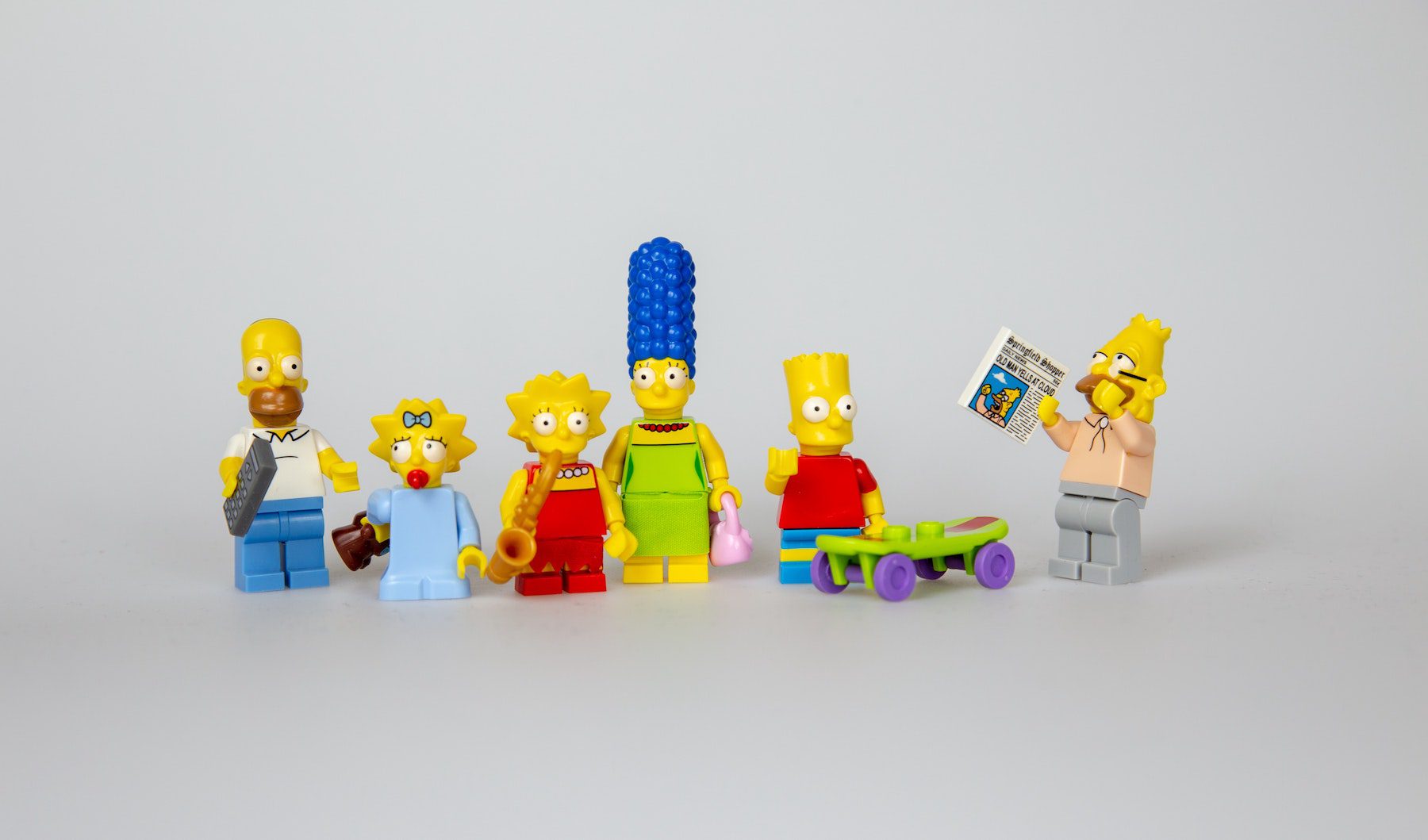 Lego minifigs of the Simpsons family. From left to right: Homer, Maggie, Lisa, Marge, Bart and his skateboard, and Grandpa Abraham Simpson holding a newspaper and yelling at the sky.