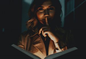 Blonde woman reading a horror novel with finger held to her lips and bathed in eerie light from below.