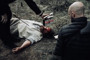 Horror movie set of a murdered man lying bloody and unconscious while a man holds a clapperboard to start the scene.