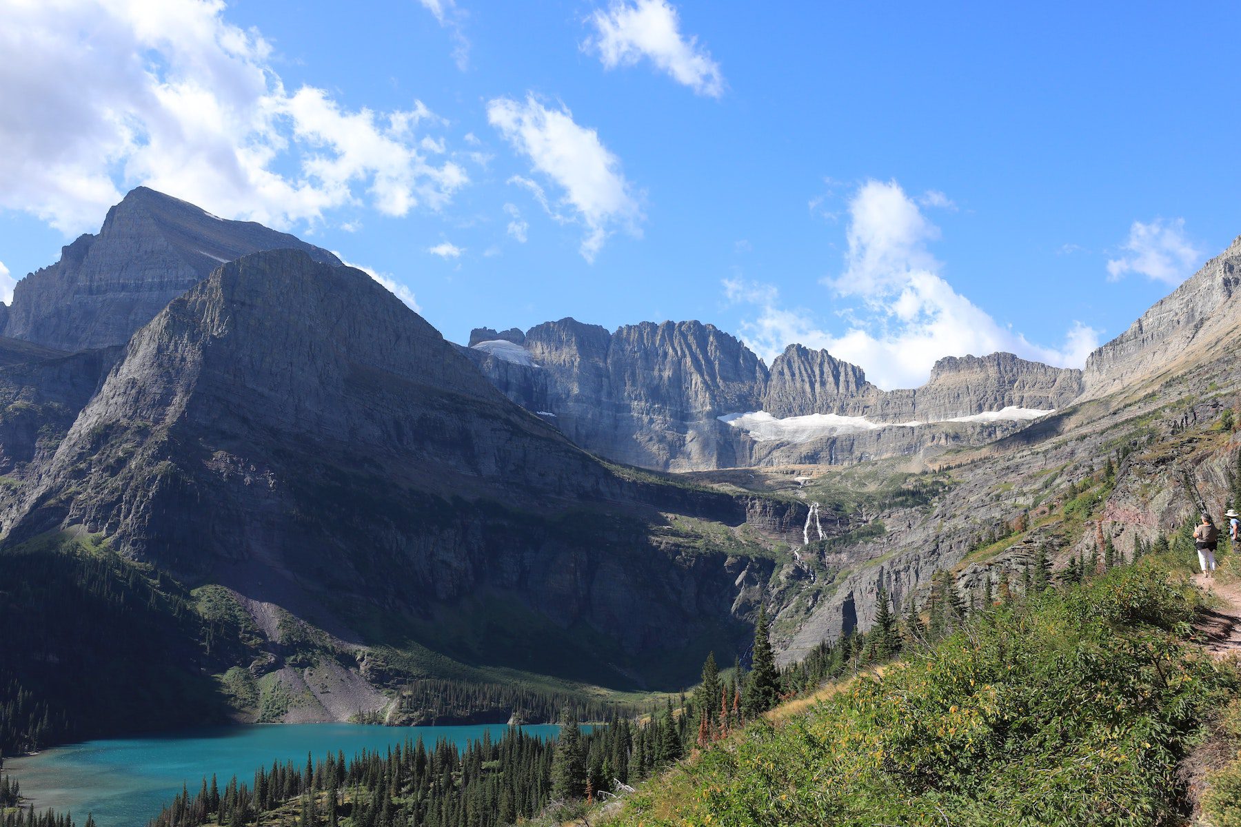 Grinnell Lake at Glacier National Park depicting a tree-lined hill with soaring mountains behind the lake