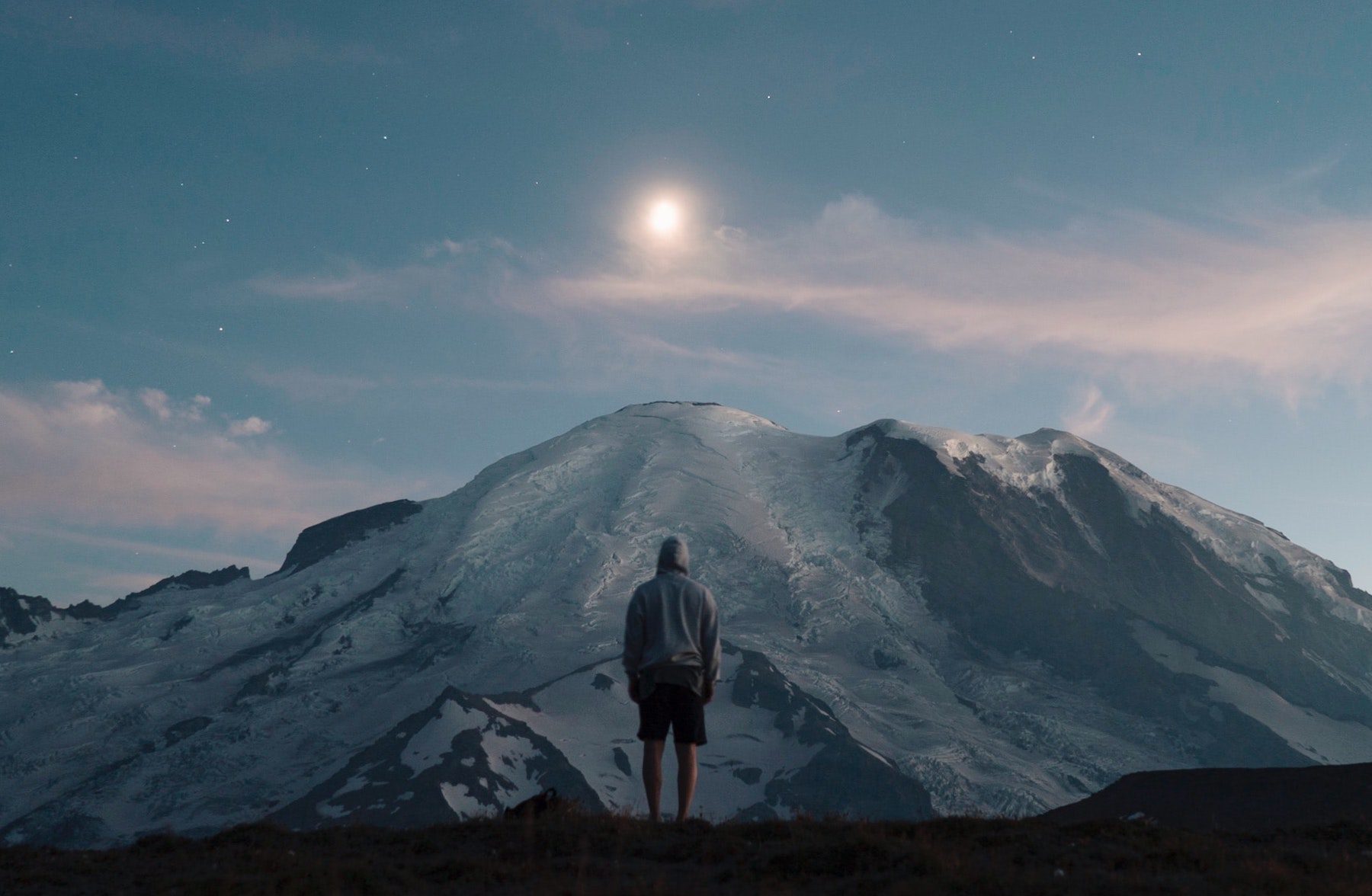 Lone hiker watching moonrise against a starry sky over a snowy peak at Mount Rainier.