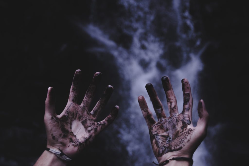 Upturned hands covered in mud or dark substance with a black and grey smoky background. Photo by Stormseeker on Unsplash